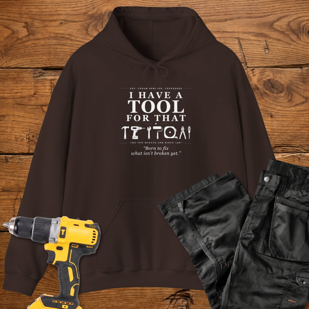 A dark chocolate coloured pullover hoodie laid flat on a rustic wooden surface, printed with the phrase 'I HAVE A TOOL FOR THAT' above a row of tool icons and smaller text quote 'Born to fix what isn't broken yet' . A yellow cordless drill and black work trousers are placed around it as styling props.