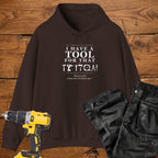 A dark chocolate coloured pullover hoodie laid flat on a rustic wooden surface, printed with the phrase 'I HAVE A TOOL FOR THAT' above a row of tool icons and smaller text quote 'Born to fix what isn't broken yet' . A yellow cordless drill and black work trousers are placed around it as styling props.