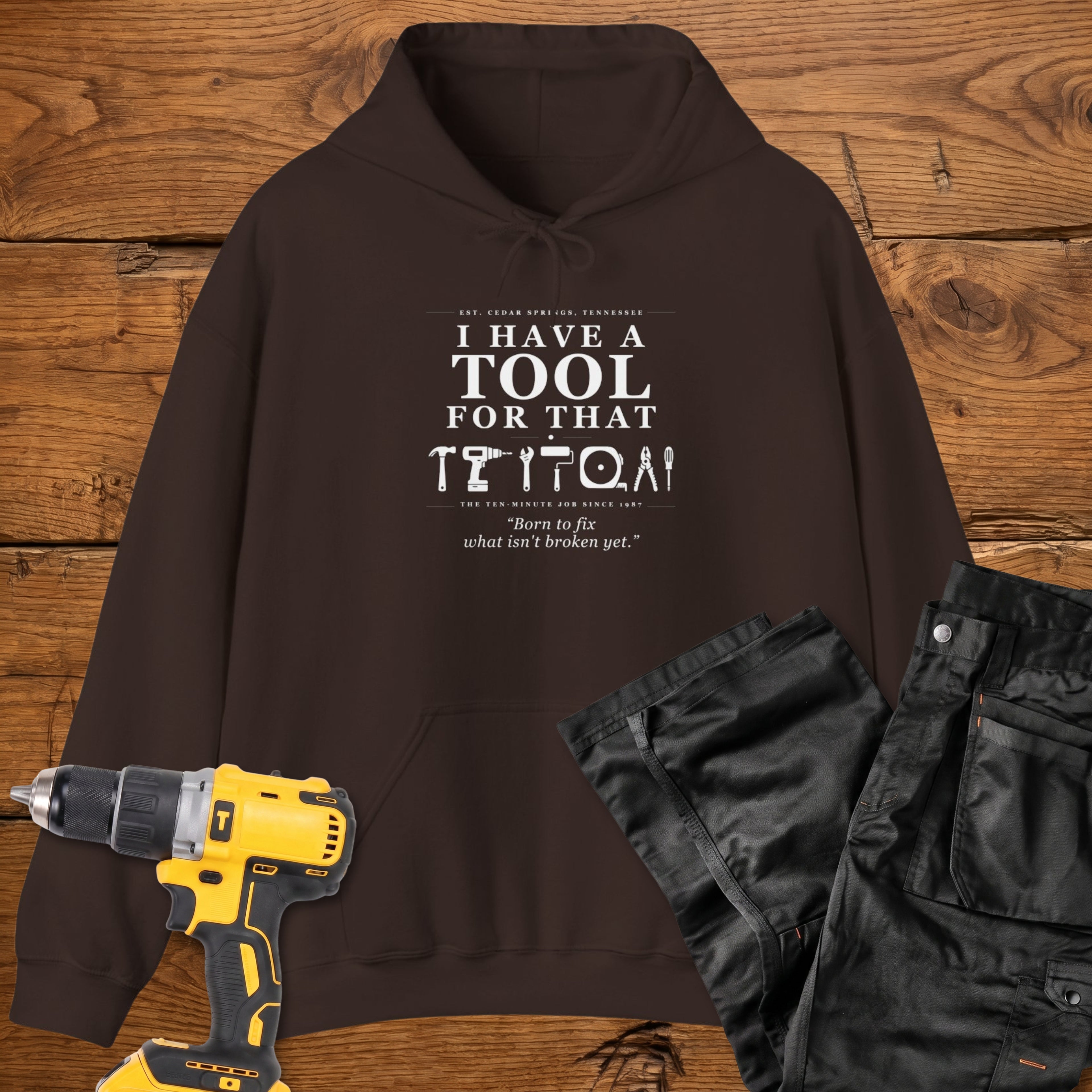 A dark chocolate coloured pullover hoodie laid flat on a rustic wooden surface, printed with the phrase 'I HAVE A TOOL FOR THAT' above a row of tool icons and smaller text quote 'Born to fix what isn't broken yet' . A yellow cordless drill and black work trousers are placed around it as styling props.