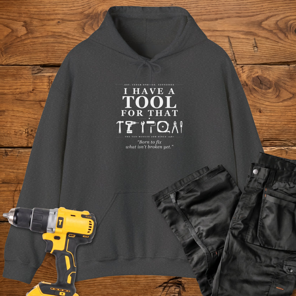 A dark heather coloured pullover hoodie laid flat on a rustic wooden surface, printed with the phrase 'I HAVE A TOOL FOR THAT' above a row of tool icons and smaller text quote 'Born to fix what isn't broken yet' . A yellow cordless drill and black work trousers are placed around it as styling props.
