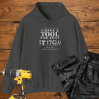 A dark heather coloured pullover hoodie laid flat on a rustic wooden surface, printed with the phrase 'I HAVE A TOOL FOR THAT' above a row of tool icons and smaller text quote 'Born to fix what isn't broken yet' . A yellow cordless drill and black work trousers are placed around it as styling props.
