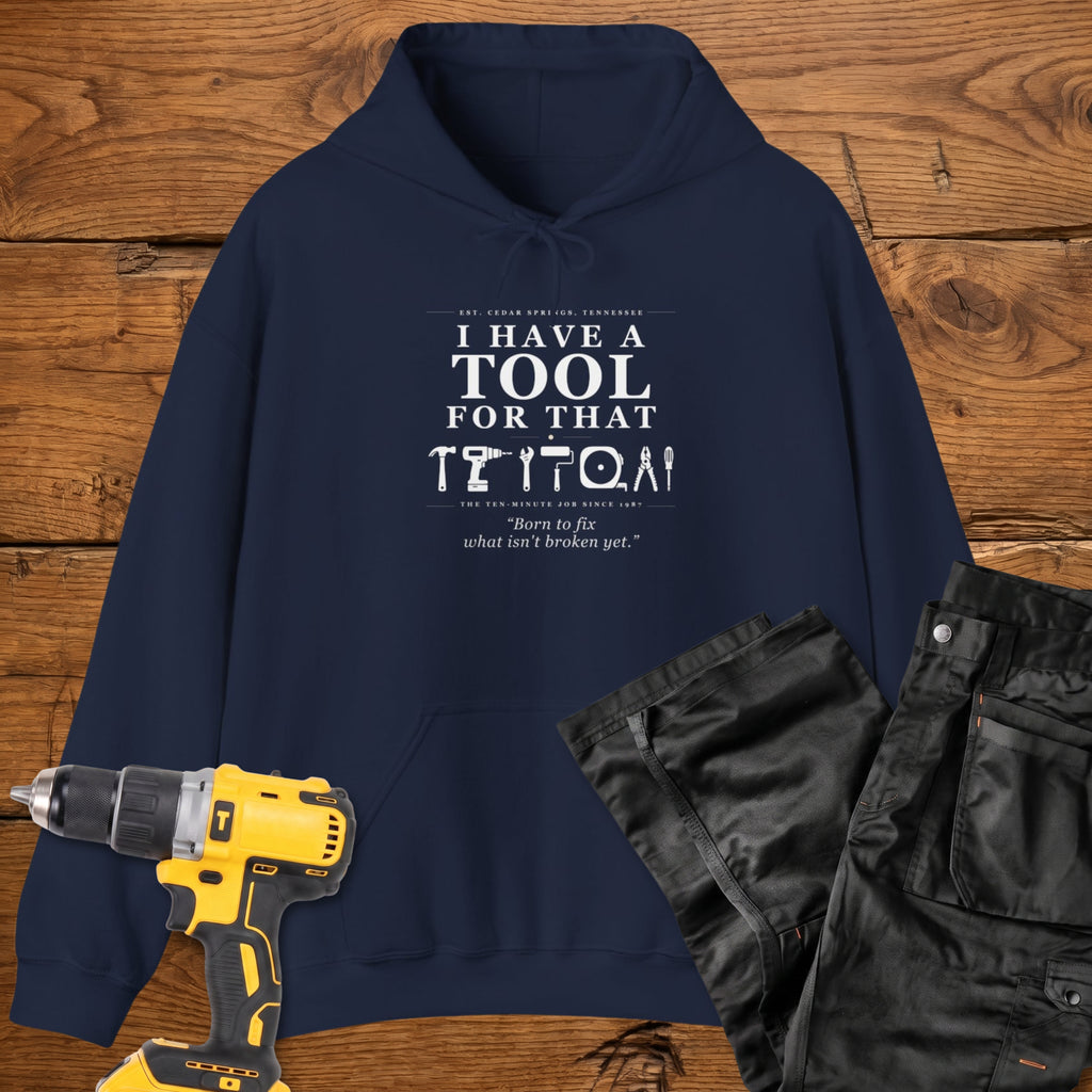 A navy coloured pullover hoodie laid flat on a rustic wooden surface, printed with the phrase 'I HAVE A TOOL FOR THAT' above a row of tool icons and smaller text quote 'Born to fix what isn't broken yet' . A yellow cordless drill and black work trousers are placed around it as styling props.