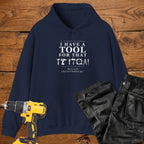 A navy coloured pullover hoodie laid flat on a rustic wooden surface, printed with the phrase 'I HAVE A TOOL FOR THAT' above a row of tool icons and smaller text quote 'Born to fix what isn't broken yet' . A yellow cordless drill and black work trousers are placed around it as styling props.