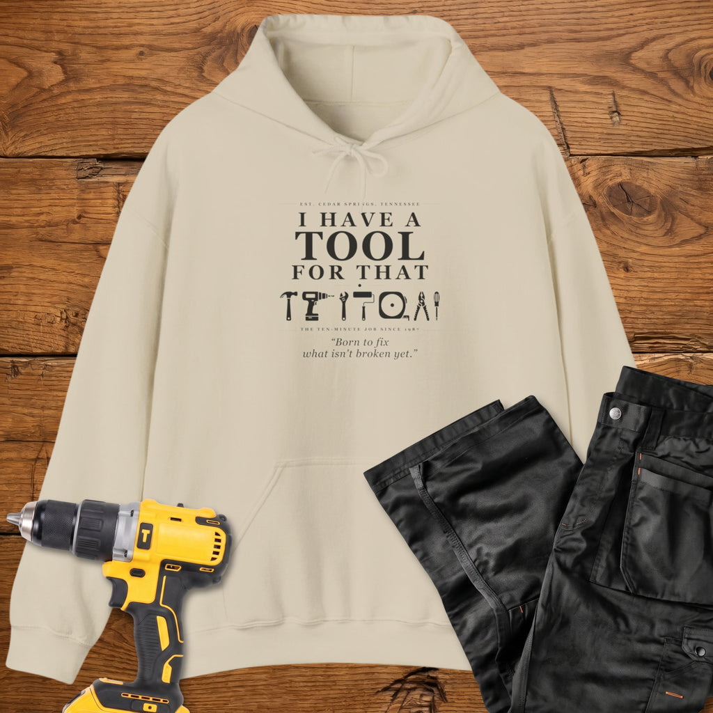 A sand coloured pullover hoodie laid flat on a rustic wooden surface, printed with the phrase 'I HAVE A TOOL FOR THAT' above a row of tool icons and smaller text quote 'Born to fix what isn't broken yet' . A yellow cordless drill and black work trousers are placed around it as styling props.
