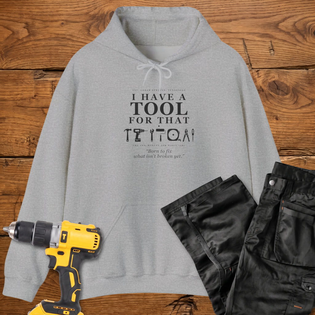 A sport grey coloured pullover hoodie laid flat on a rustic wooden surface, printed with the phrase 'I HAVE A TOOL FOR THAT' above a row of tool icons and smaller text quote 'Born to fix what isn't broken yet' . A yellow cordless drill and black work trousers are placed around it as styling props.