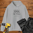 A sport grey coloured pullover hoodie laid flat on a rustic wooden surface, printed with the phrase 'I HAVE A TOOL FOR THAT' above a row of tool icons and smaller text quote 'Born to fix what isn't broken yet' . A yellow cordless drill and black work trousers are placed around it as styling props.