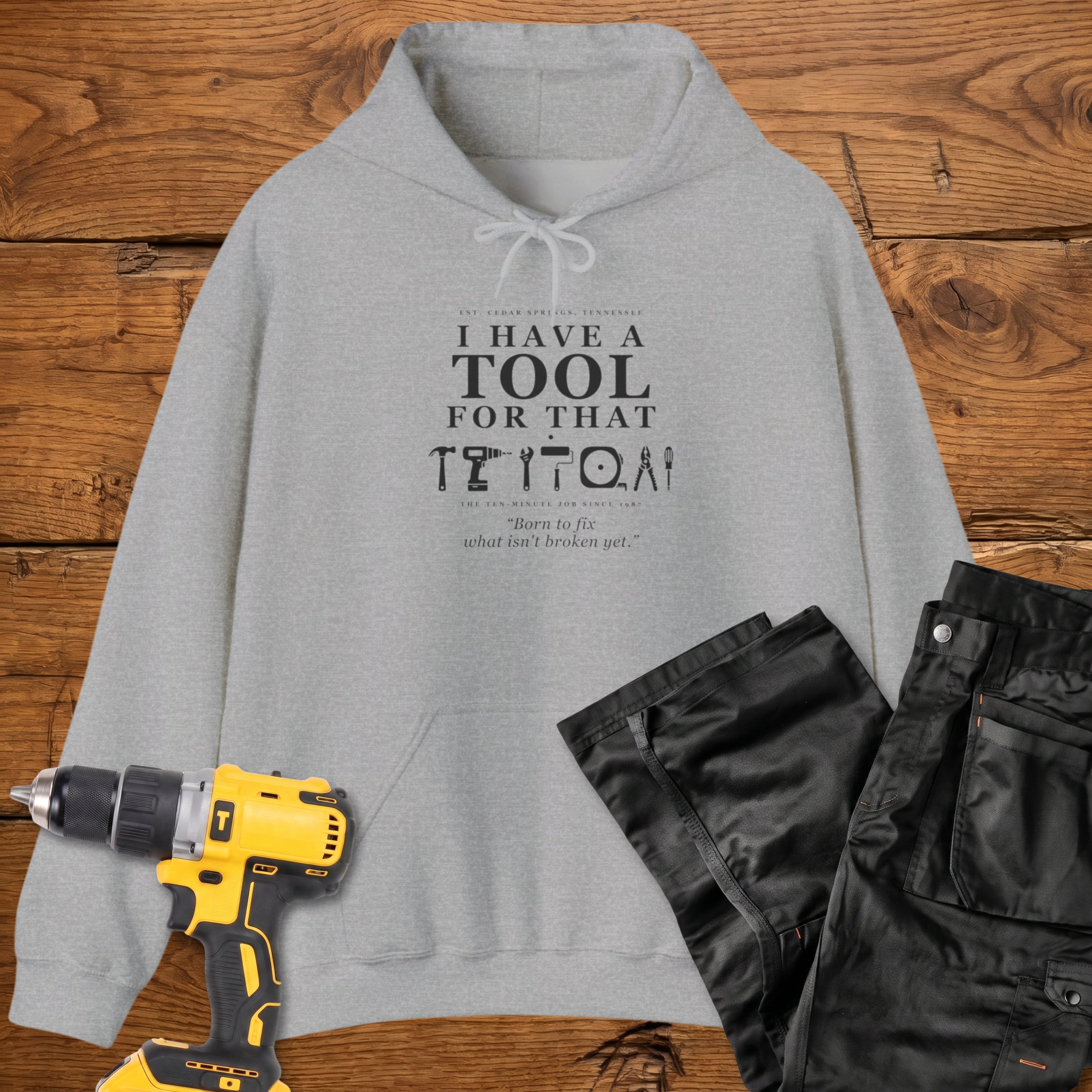 A sport grey coloured pullover hoodie laid flat on a rustic wooden surface, printed with the phrase 'I HAVE A TOOL FOR THAT' above a row of tool icons and smaller text quote 'Born to fix what isn't broken yet' . A yellow cordless drill and black work trousers are placed around it as styling props.