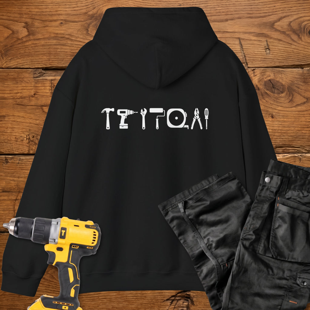 A black coloured hoodie laid flat on a rustic wooden surface, featuring a white tool silhouette across the back. A yellow cordless drill and black work trousers are placed beside it as styling props.