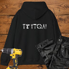 A black coloured hoodie laid flat on a rustic wooden surface, featuring a white tool silhouette across the back. A yellow cordless drill and black work trousers are placed beside it as styling props.