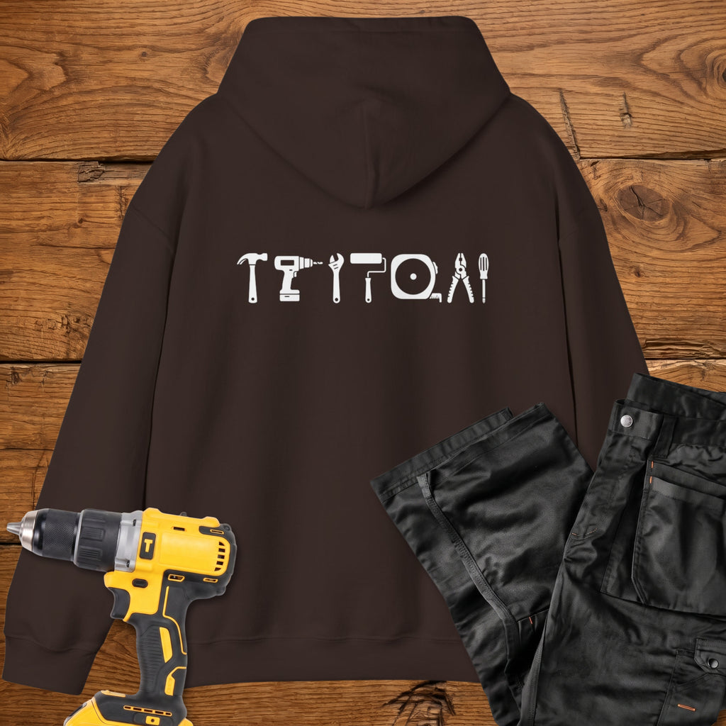 A dark chocolate coloured hoodie laid flat on a rustic wooden surface, featuring a white tool silhouette across the back. A yellow cordless drill and black work trousers are placed beside it as styling props.
