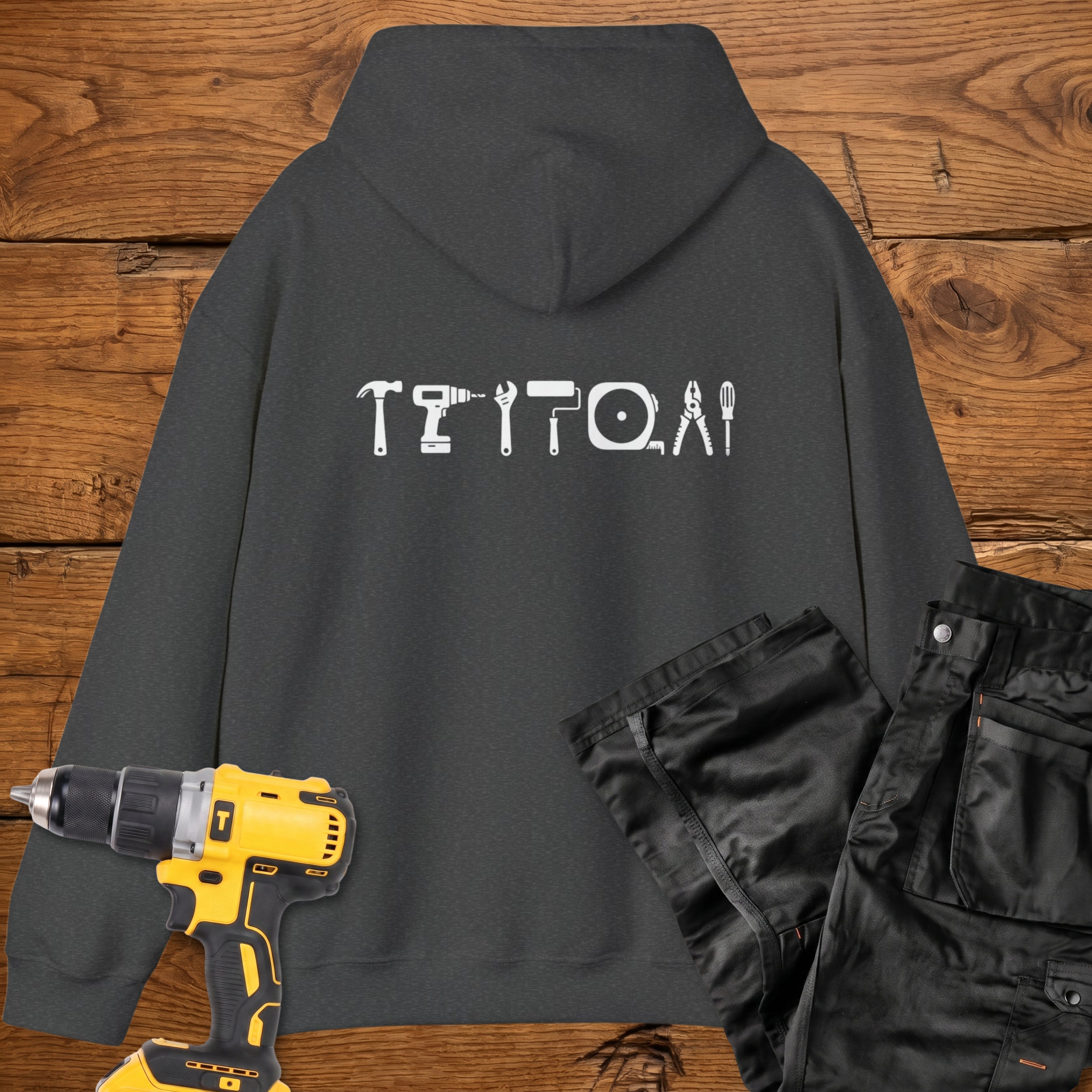 A dark heather coloured hoodie laid flat on a rustic wooden surface, featuring a white tool silhouette across the back. A yellow cordless drill and black work trousers are placed beside it as styling props.