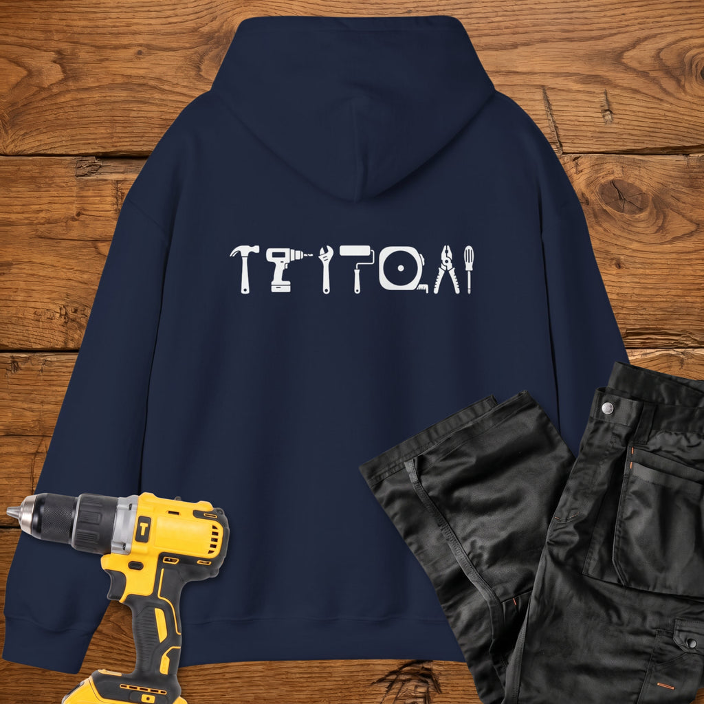 A navy coloured hoodie laid flat on a rustic wooden surface, featuring a white tool silhouette across the back. A yellow cordless drill and black work trousers are placed beside it as styling props.