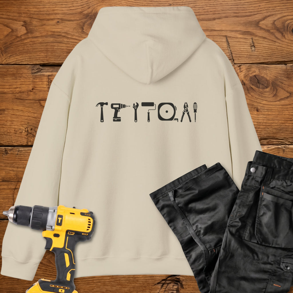 A sand coloured hoodie laid flat on a rustic wooden surface, featuring a white tool silhouette across the back. A yellow cordless drill and black work trousers are placed beside it as styling props.
