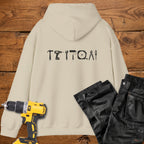 A sand coloured hoodie laid flat on a rustic wooden surface, featuring a white tool silhouette across the back. A yellow cordless drill and black work trousers are placed beside it as styling props.