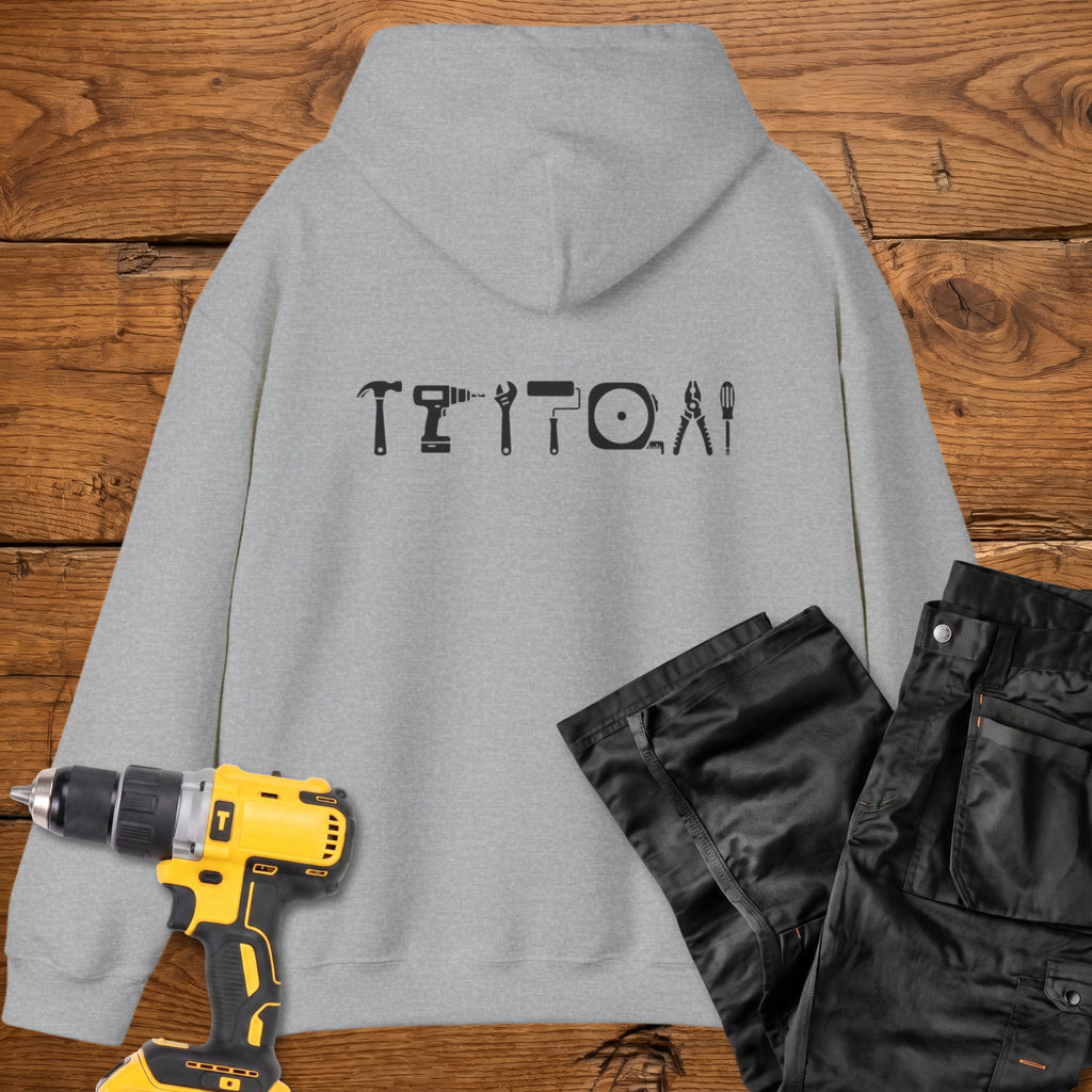 A sport grey coloured hoodie laid flat on a rustic wooden surface, featuring a white tool silhouette across the back. A yellow cordless drill and black work trousers are placed beside it as styling props.