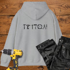 A sport grey coloured hoodie laid flat on a rustic wooden surface, featuring a white tool silhouette across the back. A yellow cordless drill and black work trousers are placed beside it as styling props.