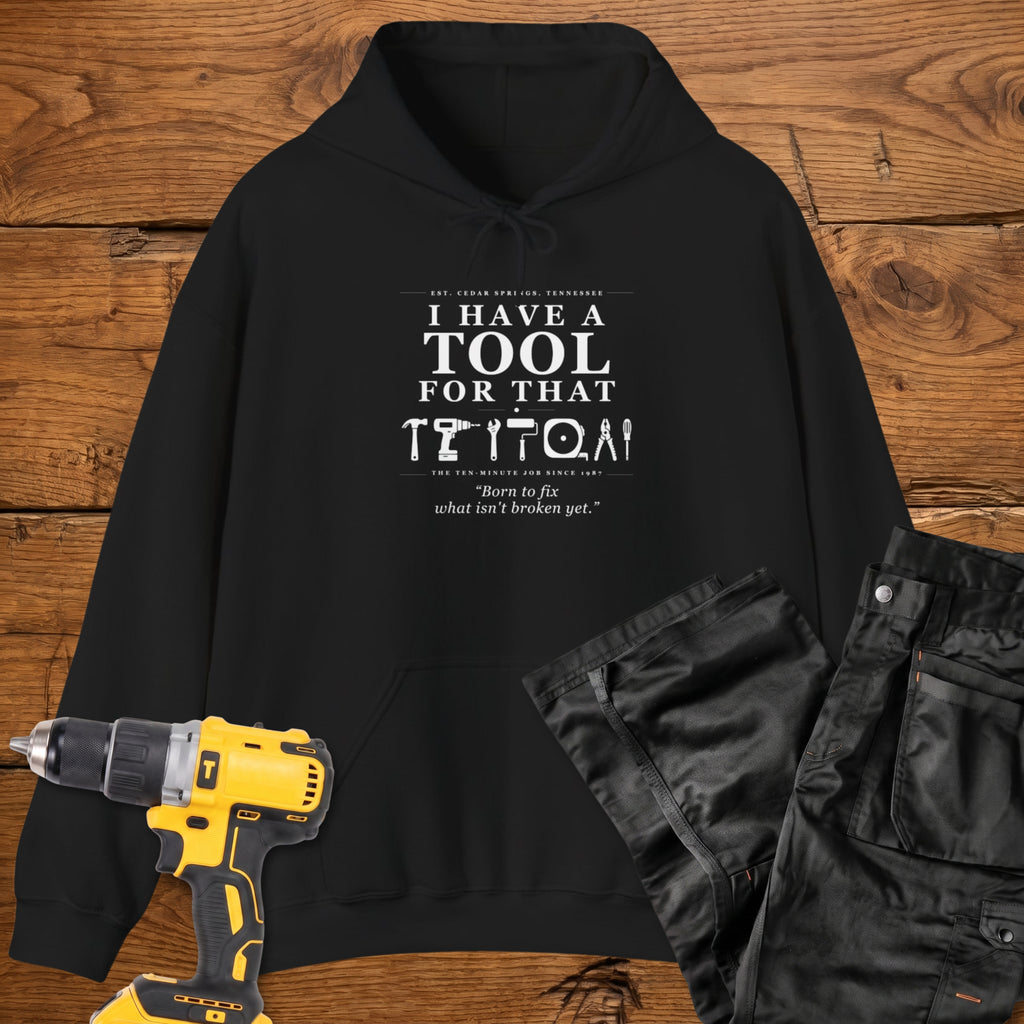 A black coloured pullover hoodie laid flat on a rustic wooden surface, printed with the phrase 'I HAVE A TOOL FOR THAT' above a row of tool icons and smaller text quote 'Born to fix what isn't broken yet' . A yellow cordless drill and black work trousers are placed around it as styling props.