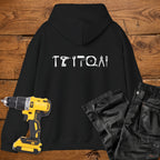 A black coloured hoodie laid flat on a rustic wooden surface, featuring a white tool silhouette across the back. A yellow cordless drill and black work trousers are placed beside it as styling props.