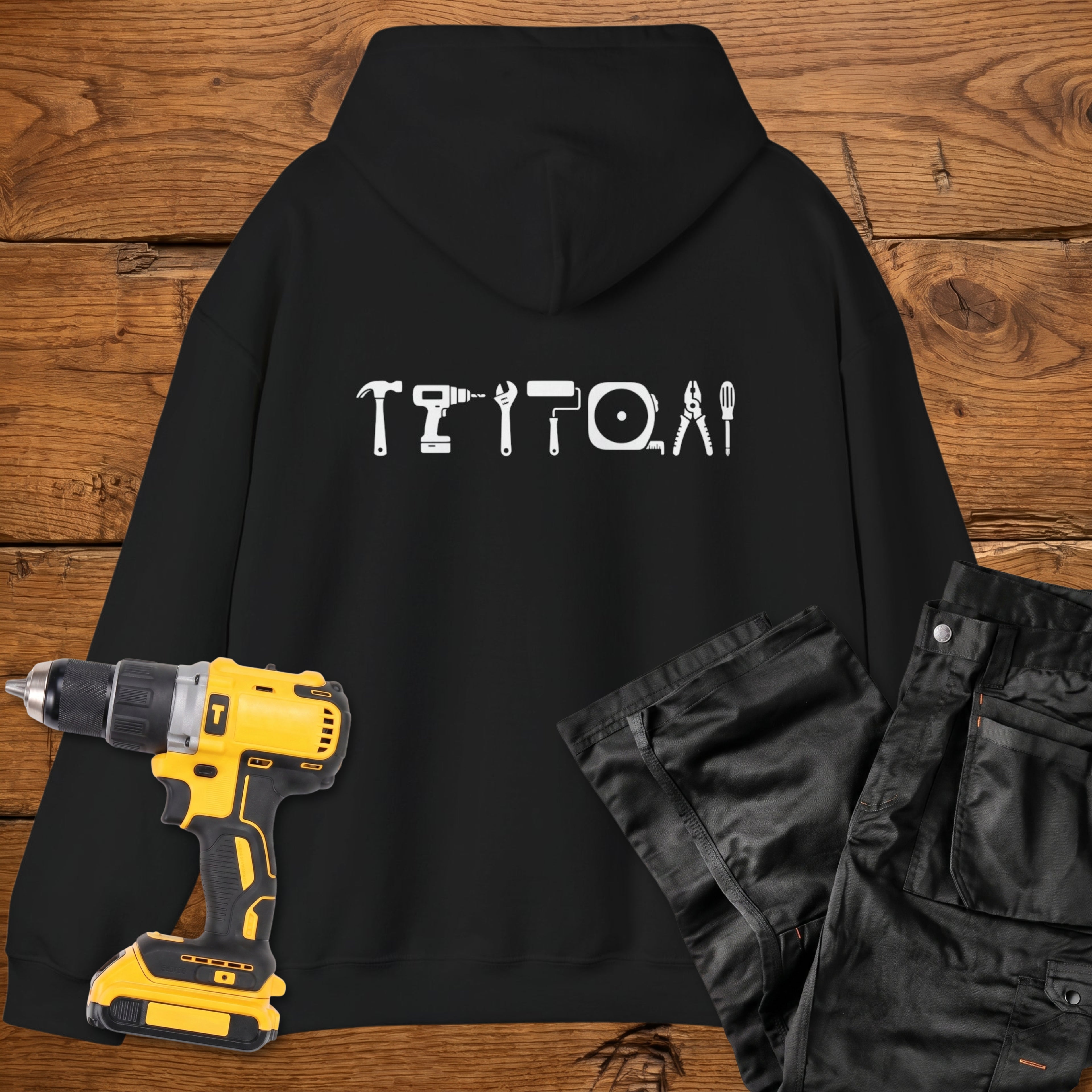 A black coloured hoodie laid flat on a rustic wooden surface, featuring a white tool silhouette across the back. A yellow cordless drill and black work trousers are placed beside it as styling props.
