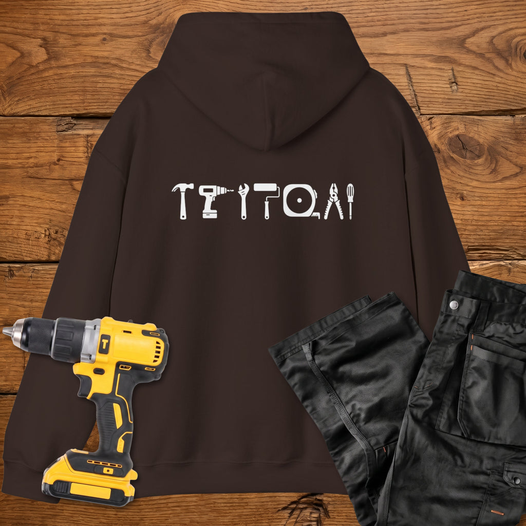 A dark chocolate coloured hoodie laid flat on a rustic wooden surface, featuring a white tool silhouette across the back. A yellow cordless drill and black work trousers are placed beside it as styling props.