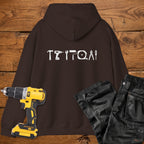 A dark chocolate coloured hoodie laid flat on a rustic wooden surface, featuring a white tool silhouette across the back. A yellow cordless drill and black work trousers are placed beside it as styling props.