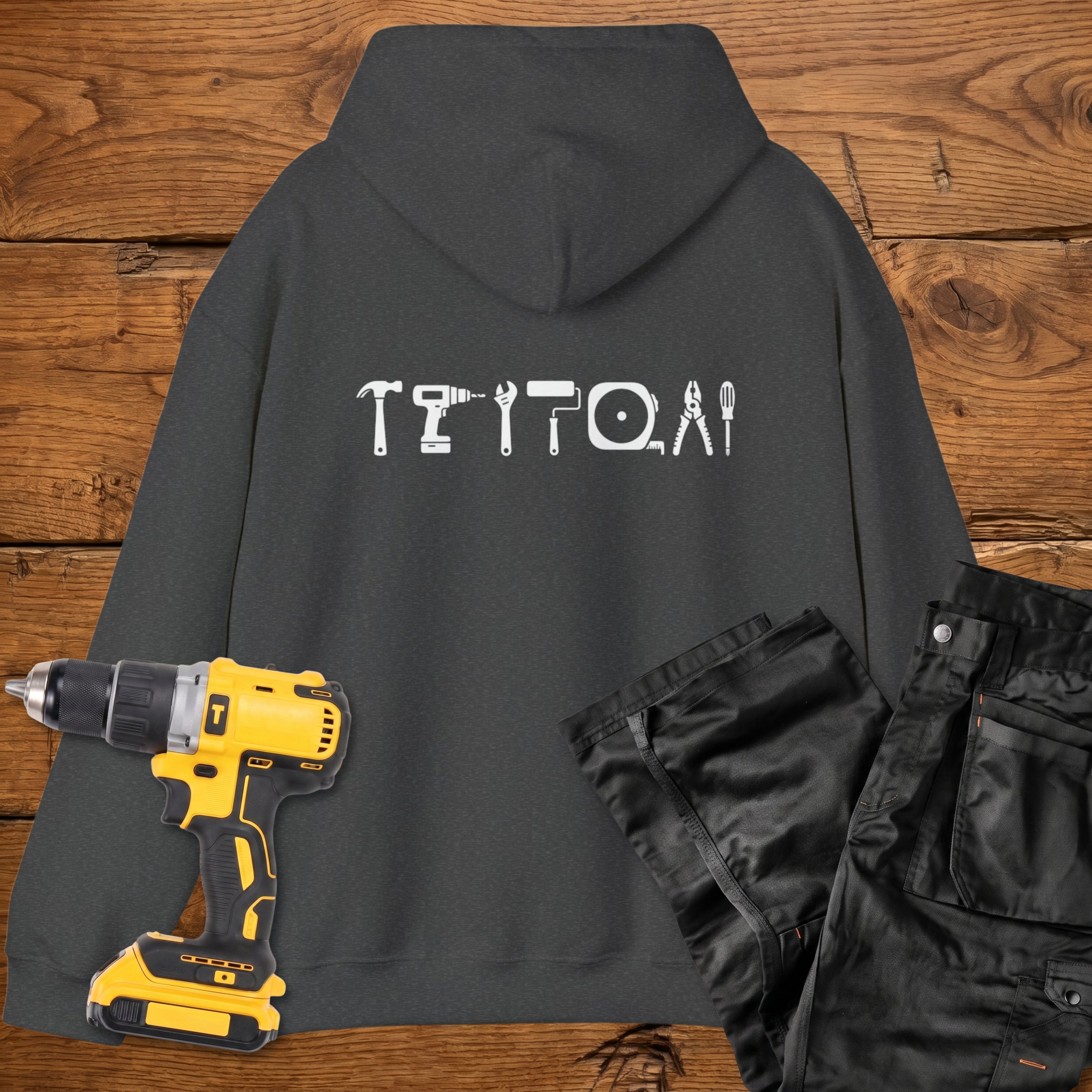 A dark heather coloured hoodie laid flat on a rustic wooden surface, featuring a white tool silhouette across the back. A yellow cordless drill and black work trousers are placed beside it as styling props.
