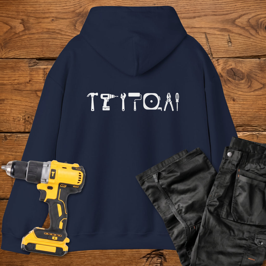 A navy blue coloured hoodie laid flat on a rustic wooden surface, featuring a white tool silhouette across the back. A yellow cordless drill and black work trousers are placed beside it as styling props.