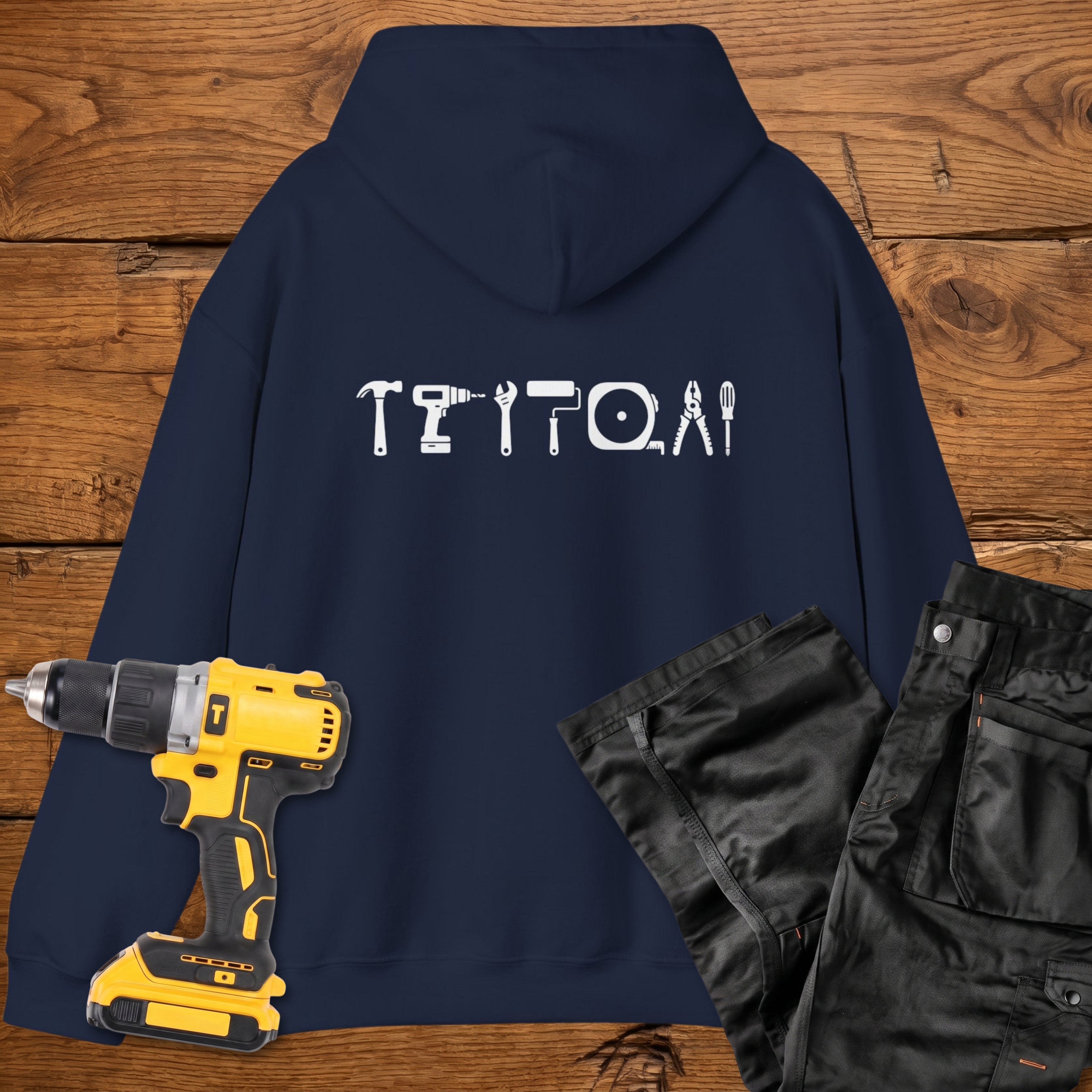 A navy blue coloured hoodie laid flat on a rustic wooden surface, featuring a white tool silhouette across the back. A yellow cordless drill and black work trousers are placed beside it as styling props.
