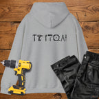 A sport grey coloured hoodie laid flat on a rustic wooden surface, featuring a white tool silhouette across the back. A yellow cordless drill and black work trousers are placed beside it as styling props.