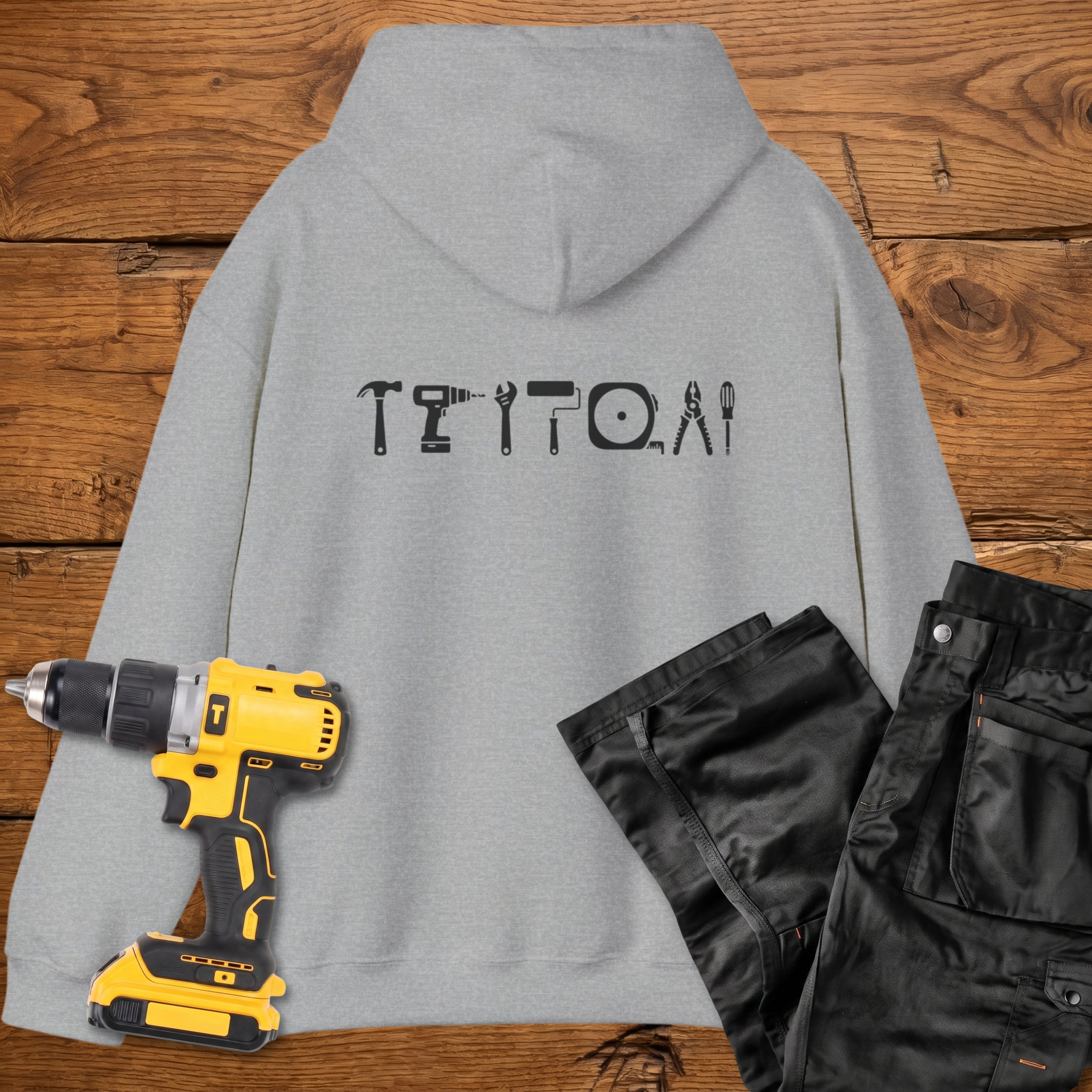 A sport grey coloured hoodie laid flat on a rustic wooden surface, featuring a white tool silhouette across the back. A yellow cordless drill and black work trousers are placed beside it as styling props.
