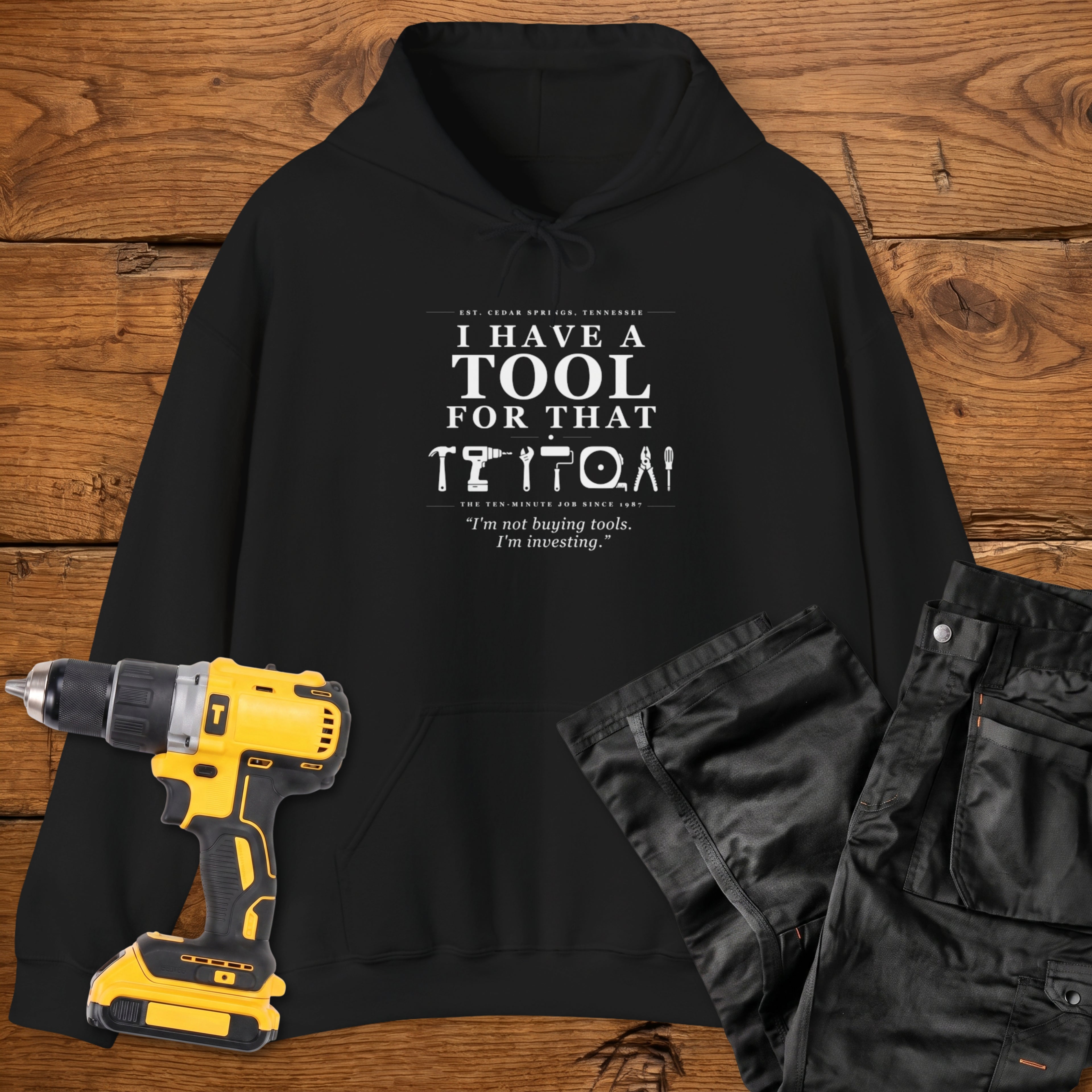 A black coloured hoodie laid flat on a wooden surface, printed with the white text 'I HAVE A TOOL FOR THAT' and a tool silhouette, plus the quote 'I'm not buying tools. I'm investing. A yellow cordless drill and black work trousers are placed beside it as styling props.