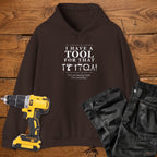 A dark chocolate coloured hoodie laid flat on a wooden surface, printed with the white text 'I HAVE A TOOL FOR THAT' and a tool silhouette, plus the quote 'I'm not buying tools. I'm investing. A yellow cordless drill and black work trousers are placed beside it as styling props.