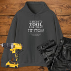 A dark heather coloured hoodie laid flat on a wooden surface, printed with the white text 'I HAVE A TOOL FOR THAT' and a tool silhouette, plus the quote 'I'm not buying tools. I'm investing. A yellow cordless drill and black work trousers are placed beside it as styling props.