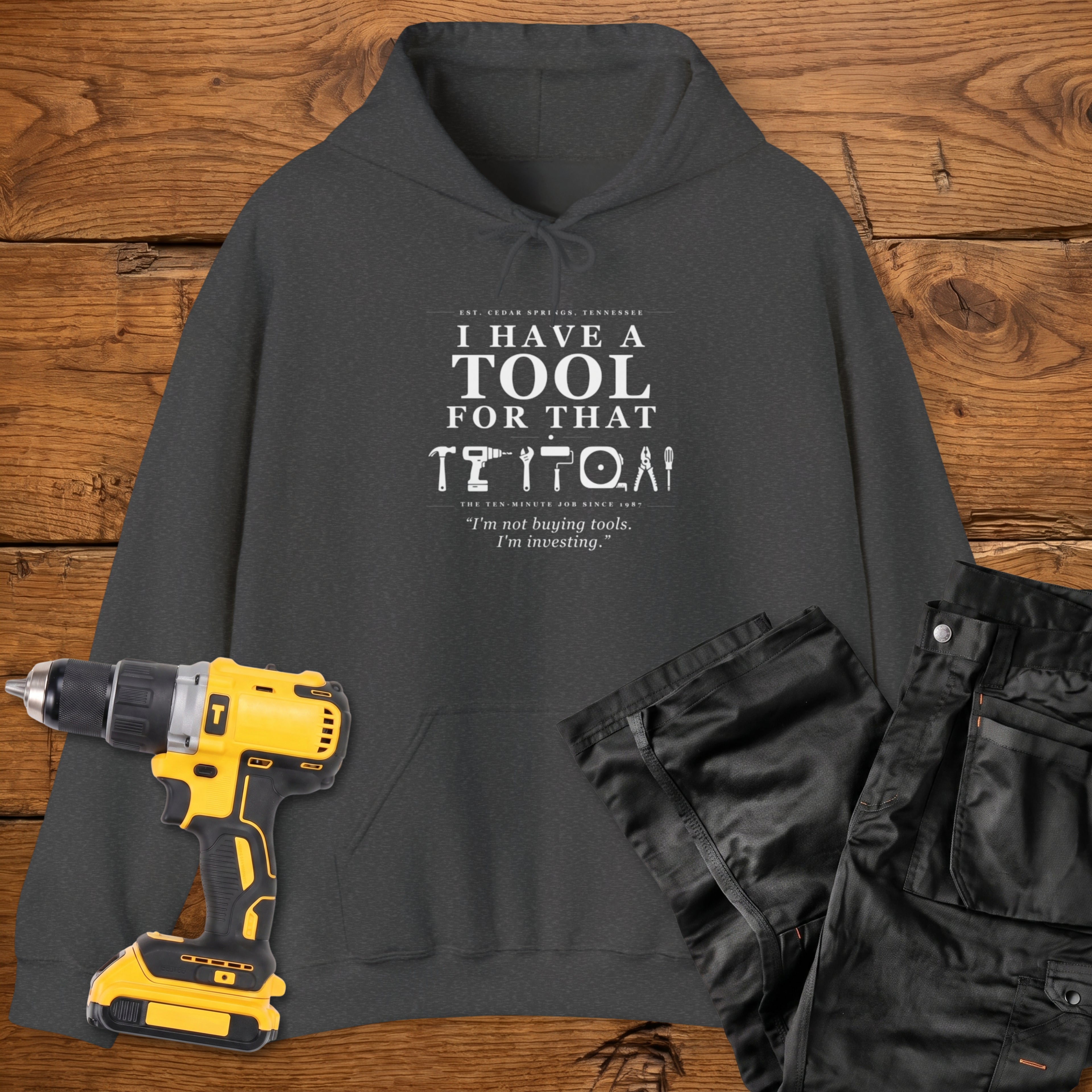 A dark heather coloured hoodie laid flat on a wooden surface, printed with the white text 'I HAVE A TOOL FOR THAT' and a tool silhouette, plus the quote 'I'm not buying tools. I'm investing. A yellow cordless drill and black work trousers are placed beside it as styling props.