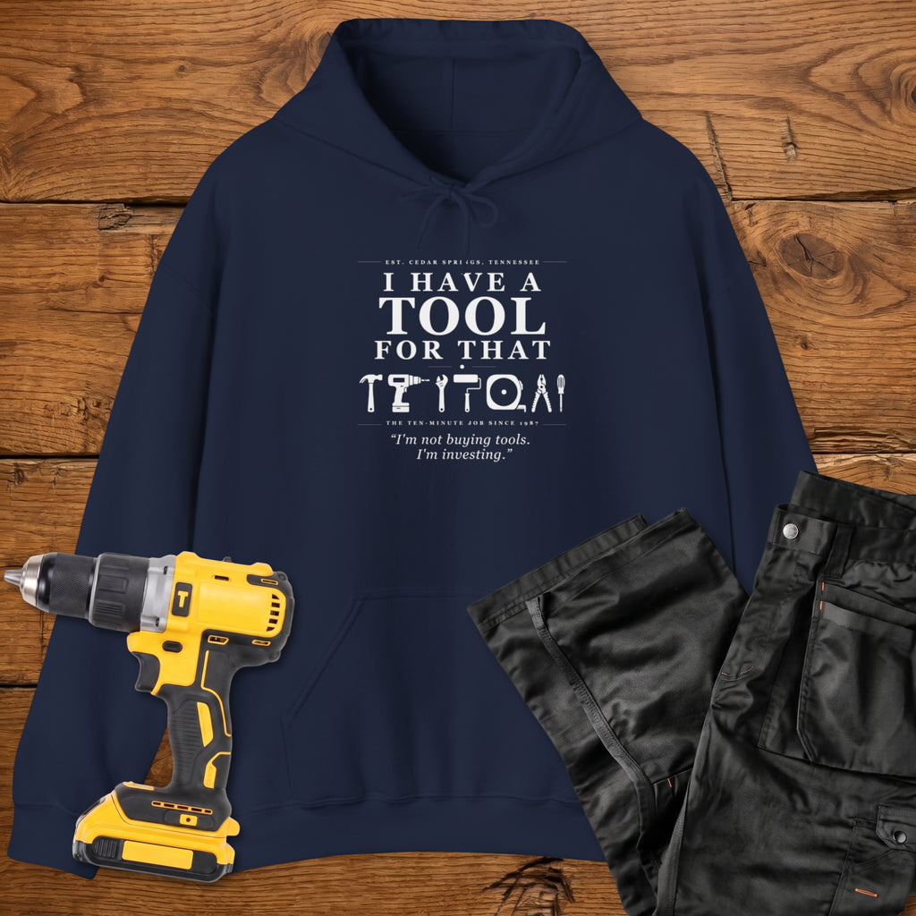A navy blue coloured hoodie laid flat on a wooden surface, printed with the white text 'I HAVE A TOOL FOR THAT' and a tool silhouette, plus the quote 'I'm not buying tools. I'm investing. A yellow cordless drill and black work trousers are placed beside it as styling props.