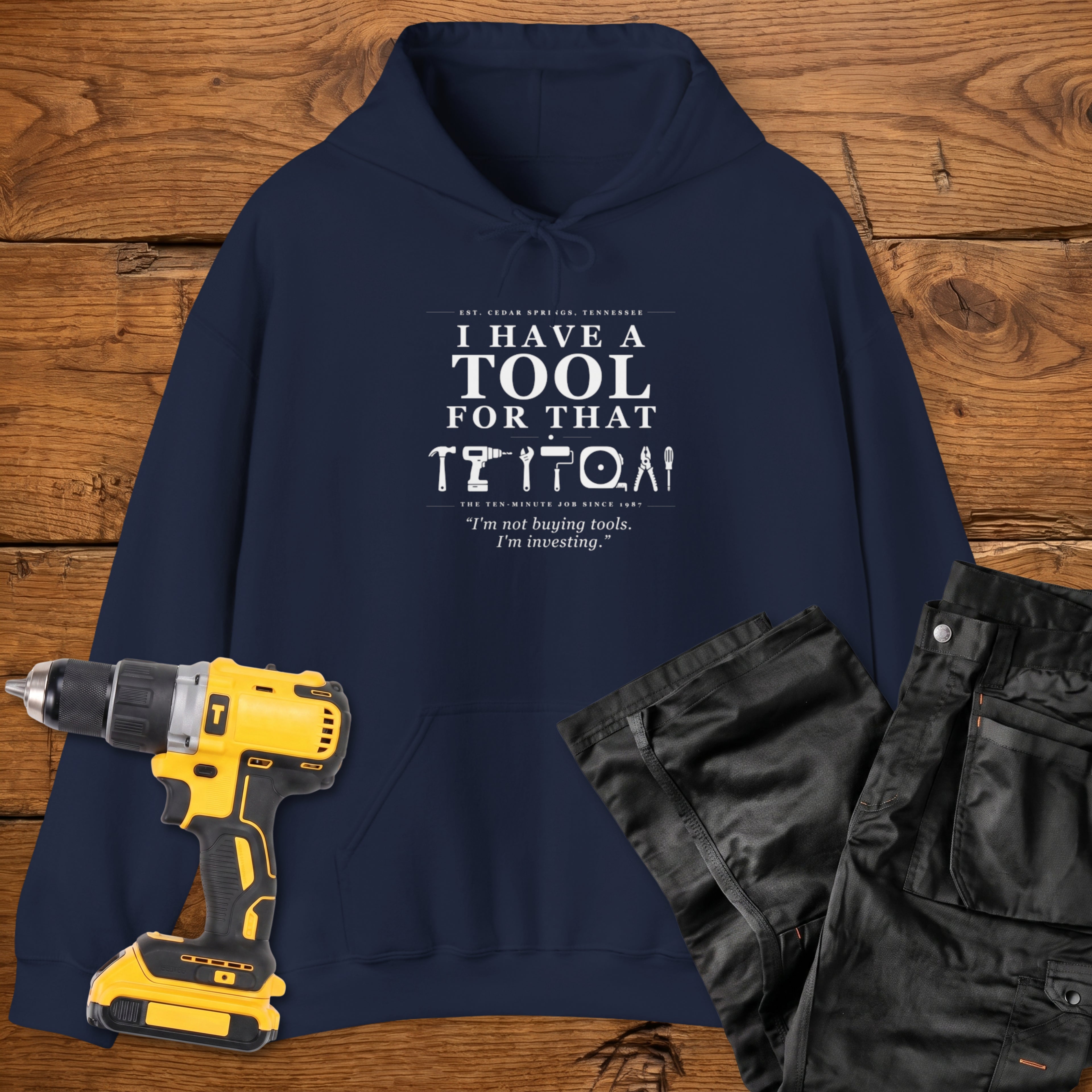 A navy blue coloured hoodie laid flat on a wooden surface, printed with the white text 'I HAVE A TOOL FOR THAT' and a tool silhouette, plus the quote 'I'm not buying tools. I'm investing. A yellow cordless drill and black work trousers are placed beside it as styling props.