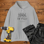 A sport grey coloured hoodie laid flat on a wooden surface, printed with the white text 'I HAVE A TOOL FOR THAT' and a tool silhouette, plus the quote 'I'm not buying tools. I'm investing. A yellow cordless drill and black work trousers are placed beside it as styling props.