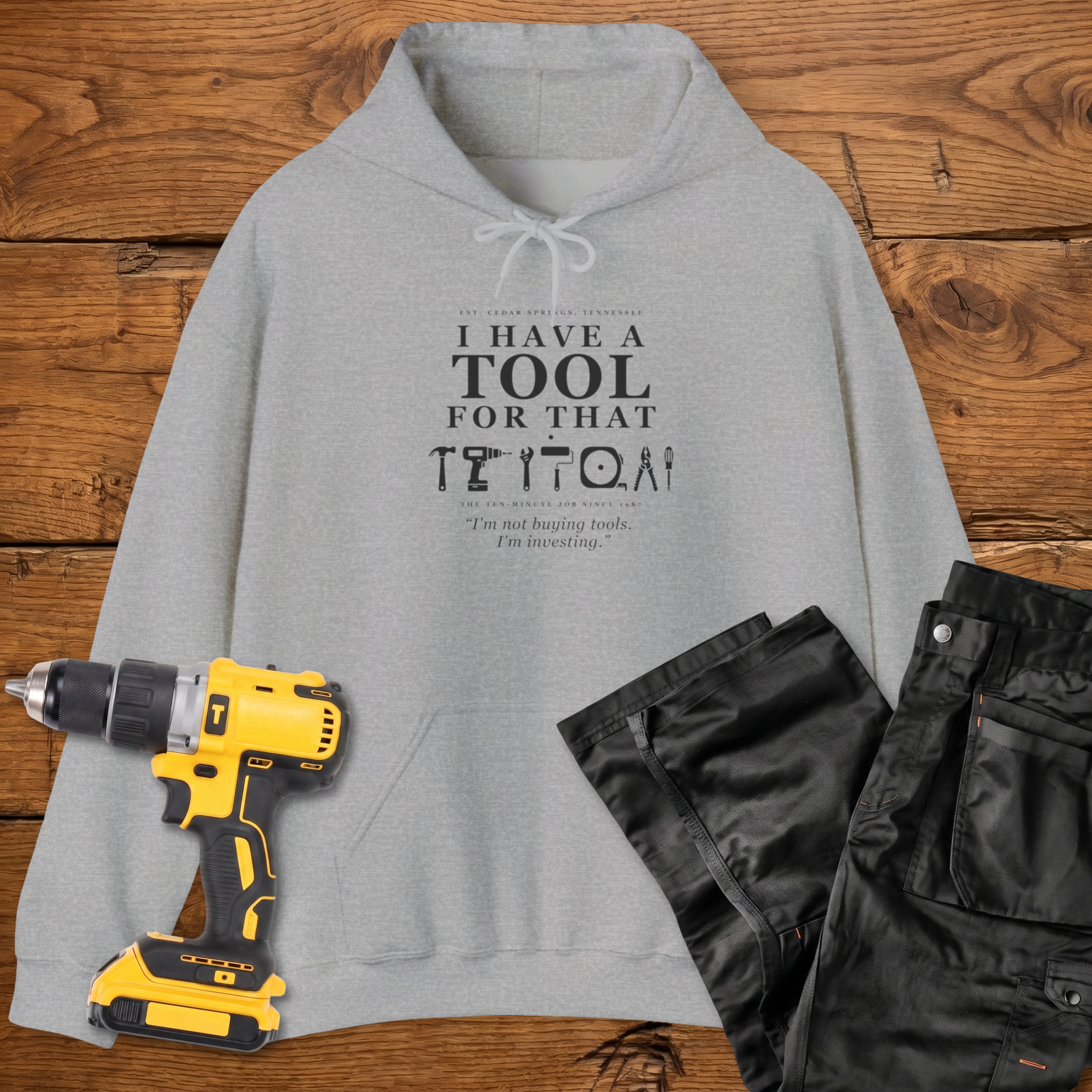 A sport grey coloured hoodie laid flat on a wooden surface, printed with the white text 'I HAVE A TOOL FOR THAT' and a tool silhouette, plus the quote 'I'm not buying tools. I'm investing. A yellow cordless drill and black work trousers are placed beside it as styling props.