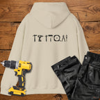 A sand coloured hoodie laid flat on a rustic wooden surface, featuring a white tool silhouette across the back. A yellow cordless drill and black work trousers are placed beside it as styling props.