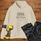 A sand coloured hoodie laid flat on a wooden surface, printed with the white text 'I HAVE A TOOL FOR THAT' and a tool silhouette, plus the quote 'I'm not buying tools. I'm investing. A yellow cordless drill and black work trousers are placed beside it as styling props.