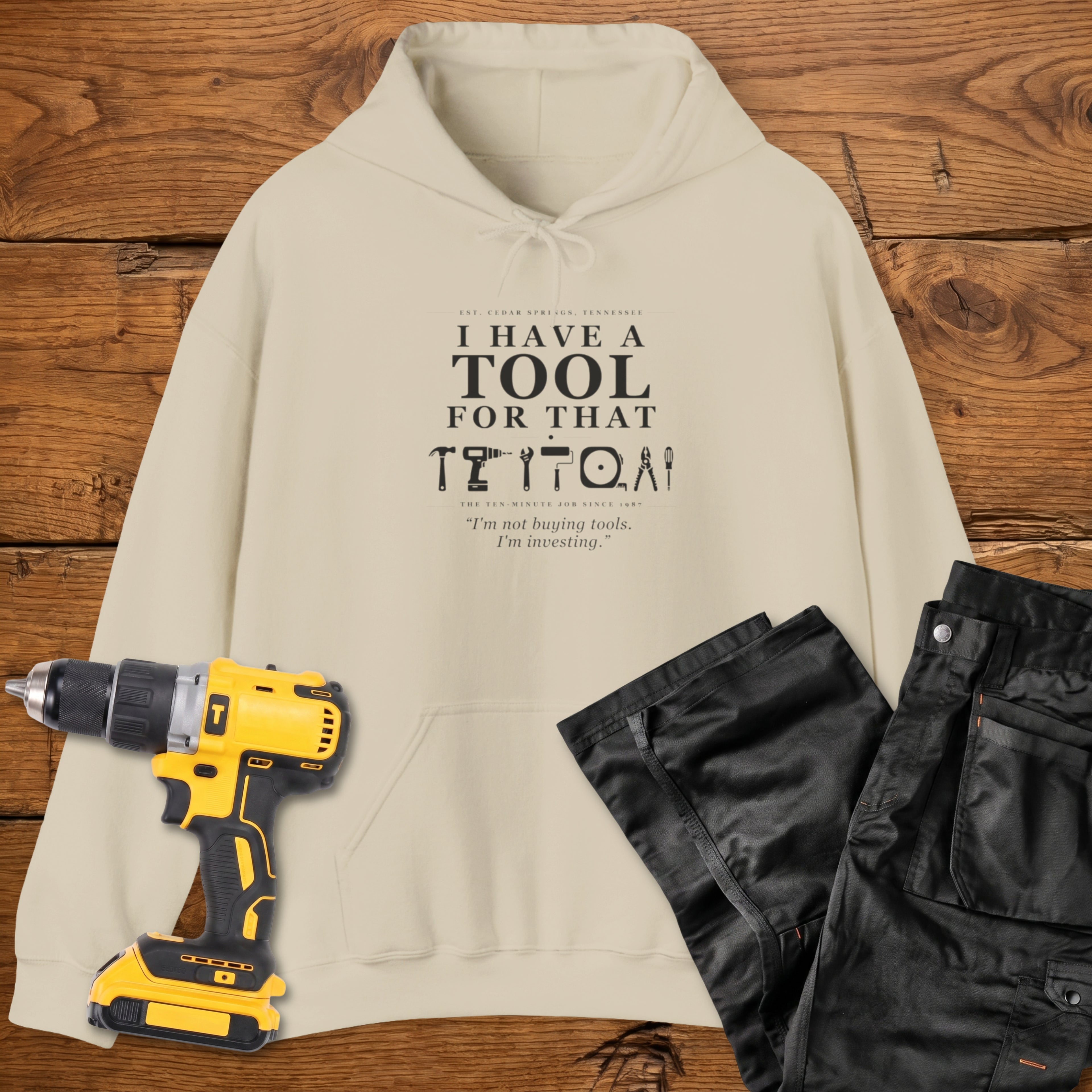 A sand coloured hoodie laid flat on a wooden surface, printed with the white text 'I HAVE A TOOL FOR THAT' and a tool silhouette, plus the quote 'I'm not buying tools. I'm investing. A yellow cordless drill and black work trousers are placed beside it as styling props.
